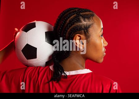 Donna afroamericana con palla da calcio sulla spalla in studio con sfondo rosso, in maglia rossa Foto Stock