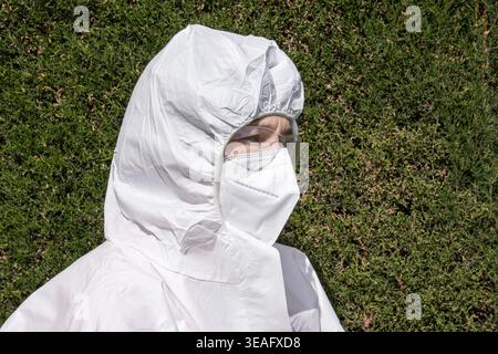 Ritratto ravvicinato di una donna che indossa una tuta bianca, maschera facciale FFP2 e occhiali protettivi su uno sfondo verde siepe. Foto Stock