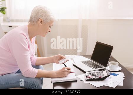 Una persona anziana lavora su documenti finanziari mentre utilizza un notebook e una calcolatrice a casa Foto Stock