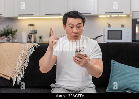 L'uomo di ispirazione asiatica a casa studia il corso online su smartphone indossa gli auricolari ascolta la lezione e trova risposta. Il ragazzo cinese sul divano mostra un segno ok imposta l'obiettivo per la settimana salva l'attività sui sorrisi dell'app inizia il piano Foto Stock