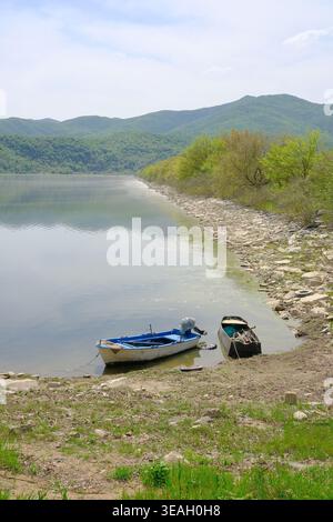 Un tranquillo paesaggio naturale caratterizzato da due piccole barche da pesca ormeggiate vicino alla riva rocciosa di un lago calmo, con dolci colline verdi sullo sfondo e. Foto Stock