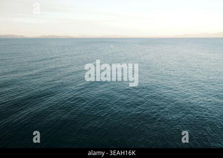 Il mare blu profondo si estende fino all'orizzonte, riflettendo la luce soffusa di un cielo pallido sopra forme di terra lontane e smorzate Foto Stock