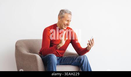 Uomo anziano sorridente e saluta durante la videochiamata mentre è seduto in sedia con sfondo semplice Foto Stock