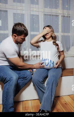 Donna incinta e partner che toccano la pancia, padre incinta che mette la mano su scritte IN MAMMA dipinte, seduta sul banco della finestra, tenda morbida sullo sfondo, caldo Foto Stock