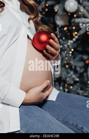 Donna incinta che tiene in mano l'ornamento di Natale rosso alla pancia, madre bianca che si aspetta che celebra le festività con l'albero festivo sullo sfondo accogliente Foto Stock