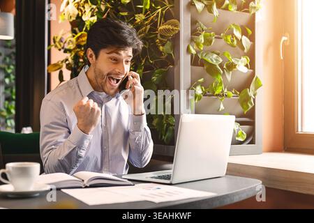 Emozionato millenaria uomo d affari con computer portatile aperto parlando al telefono Foto Stock