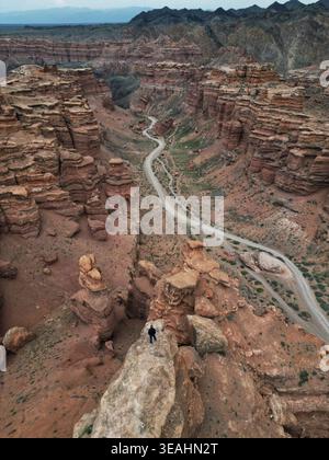Vista aerea del Black Canyon con una strada tortuosa attraverso aspre formazioni rocciose e una persona in piedi su un affioramento roccioso Aktogay, regione di Almaty, kazako Foto Stock