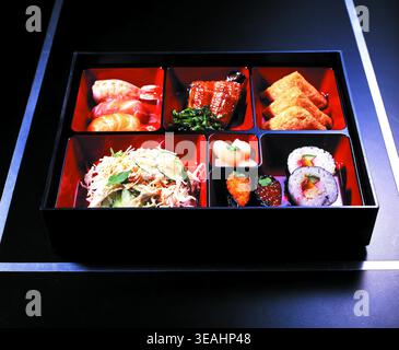 Una scatola bento nera con sei scomparti, contenente vari piatti giapponesi come sushi, insalata e anguilla. La scatola è posizionata su una superficie scura, con una sottile luce blu che la illumina dall'alto. Foto Stock
