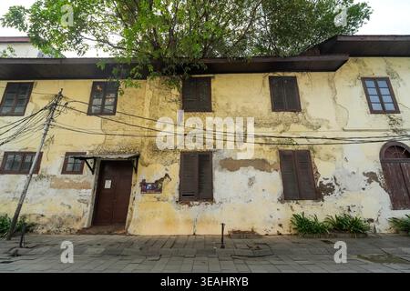Una facciata gialla intemprata di un edificio coloniale olandese nella città vecchia di Giacarta, caratterizzata da vernice sbucciante, persiane in legno e un grande albero che cresce Foto Stock