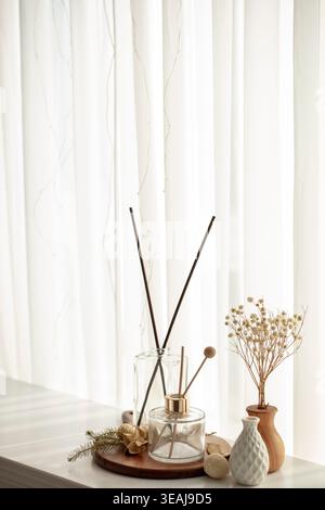 Una selezione di articoli per l'arredamento della casa, tra cui un diffusore di canne, un piccolo vaso con fiori secchi e alcuni oggetti decorativi, siedono su un davanzale davanti a una tenda bianca. Foto Stock