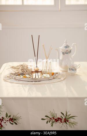 Un tavolo splendidamente allestito con un diffusore di canne, una teiera e un bouquet di fiori secchi, adagiato in una stanza soleggiata. Foto Stock