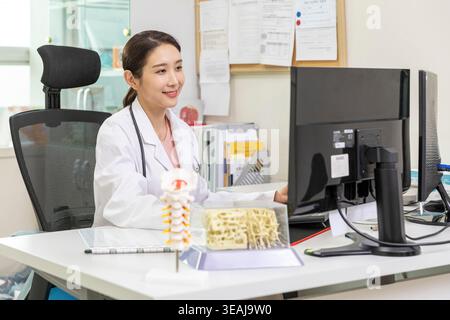 Una giovane donna asiatica si siede al suo computer in un moderno ufficio medico, rivedendo i file dei pazienti. Foto Stock
