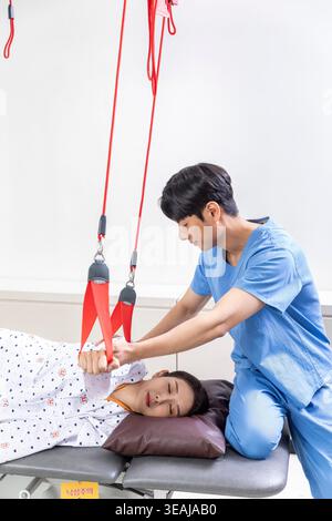 Una giovane donna in un letto d'ospedale sta ricevendo la terapia fisica da un terapeuta maschio utilizzando una cinghia di sospensione rossa per aiutarla a guarire da una lesione. Foto Stock