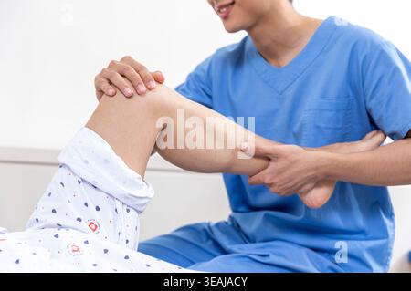 Un giovane infermiere o un medico di sesso maschile sostiene dolcemente la gamba di un paziente anziano in un letto d'ospedale. Foto Stock