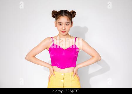 Una giovane donna con le mani sui fianchi, con un top rosa e una gonna gialla, si erge su uno sfondo neutro con una sottile ombra. Foto Stock