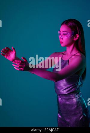 Una donna con un outfit futuristico, che indossa occhiali al neon, gesti in un ambiente buio e illuminato al neon. Foto Stock