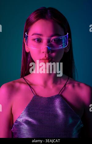 Una donna che indossa occhiali futuristici a realtà aumentata illuminati al neon, in un ambiente urbano scarsamente illuminato. Foto Stock