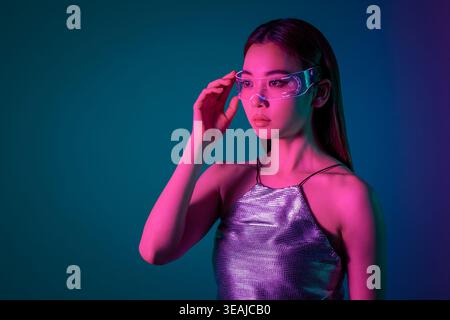 Una giovane donna che indossa occhiali futuristici per la realtà aumentata in un paesaggio urbano illuminato al neon. Foto Stock