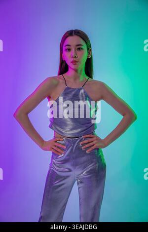Una giovane donna con i capelli lunghi sta con le mani sui fianchi e indossa un abito color argento lucido in una stanza poco illuminata e illuminata al neon. Foto Stock