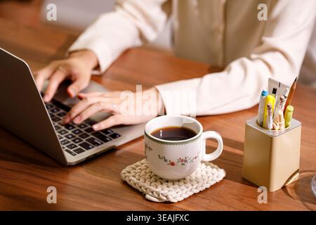 Una donna si siede a una scrivania, scrivendo sul suo computer portatile sorseggiando un caffè e circondato dalle forniture per l'ufficio. Foto Stock