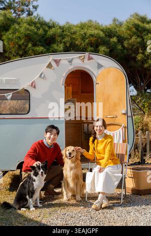 Una giovane coppia si siede di fronte a un camper d'epoca, circondato da due cani felici e da un fuoco caldo, godendosi una serata tranquilla nel bosco. Foto Stock