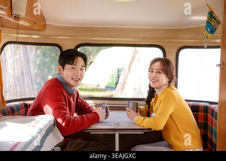 Una giovane coppia si siede insieme in un camper in stile vintage, gustando un caffè e sorridendo l'un l'altro. Foto Stock