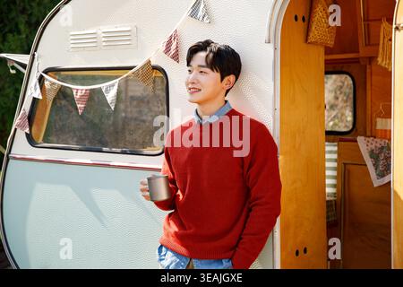 Un giovane uomo si trova di fronte a un camper vintage, tenendo in mano una tazza di caffè e sorridendo mentre si gode il sole mattutino. Foto Stock
