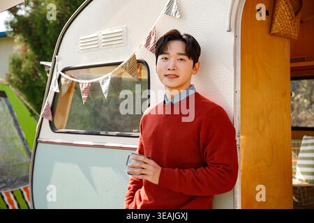 Un giovane con un maglione rosso si trova di fronte a un camper d'epoca, tenendo in mano una tazza e sorridendo davanti alla macchina fotografica. Foto Stock