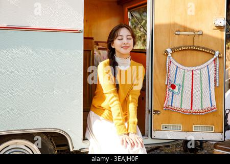 Una giovane donna con una treccia siede all'ingresso di un camper d'epoca, sorridendo e guardando il paesaggio soleggiato. Foto Stock