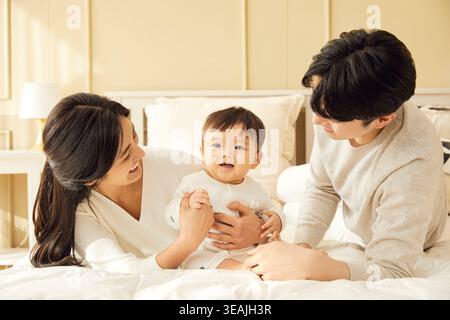 Una giovane coppia asiatica e il loro bambino giocano su un letto in una camera da letto soleggiata. Foto Stock