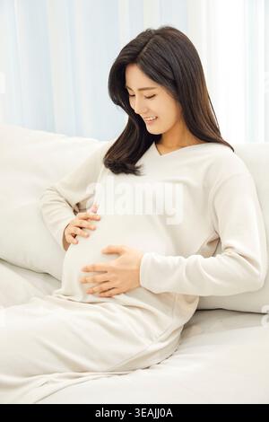 Una donna incinta siede su un divano, cullandosi dolcemente la pancia. Foto Stock