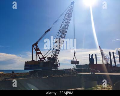 I lavoratori guidano una cassa di carico sollevata con gru sotto la luce solare intensa, catturando il coordinamento industriale e le attrezzature per impieghi gravosi Foto Stock