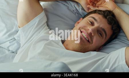 Uomo caucasico svegliarsi a letto a casa sorridente guardando la macchina fotografica felice sano maschio europeo ragazzo confortevole accogliente riposato godendo di buon giorno Foto Stock