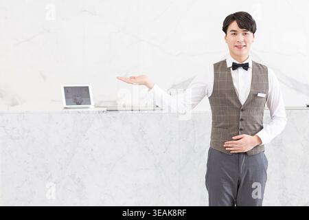 Un giovane receptionist maschile con un sorriso cordiale e un gesto di benvenuto, in piedi dietro una scrivania in marmo in una moderna lobby. Foto Stock