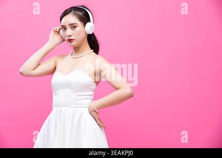 Una giovane donna con un abito da sposa bianco ascolta la musica con le sue cuffie in uno studio rosa. Foto Stock