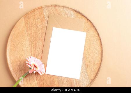 Biglietto d'auguri bianco con busta kraft e fiore di gerbera rosa su vassoio di legno Vista dall'alto composizione minima del mockup. Posizione piatta. Foto Stock