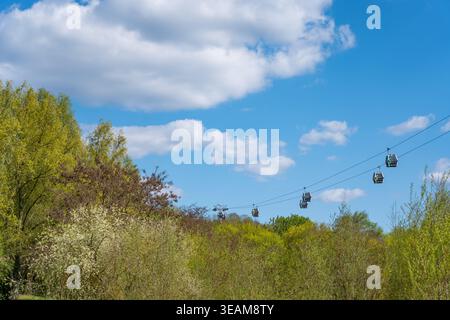 Germania Berlino 21 aprile 2026. Le funivie scivolano sopra le verdi cime degli alberi in un luminoso paesaggio primaverile con cielo blu e nuvole. Alberi in fiore e freschi Foto Stock