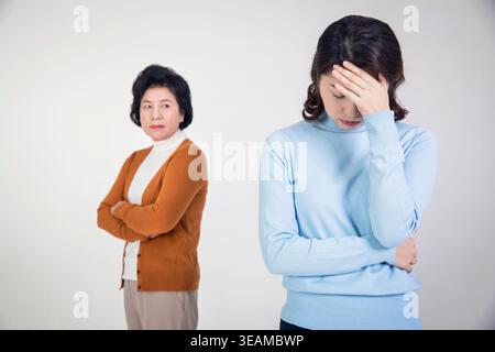 Una giovane donna tiene la testa in mano, sembrando preoccupata, mentre sua madre sta dietro di lei con le braccia incrociate, sembrando preoccupata. Foto Stock