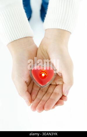 Le mani di una donna cullano una candela a forma di cuore in un supporto di vetro. Foto Stock