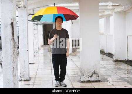 Giovane con in mano un colorato ombrello arcobaleno sotto un colonnato di cemento bianco, strada urbana Foto Stock