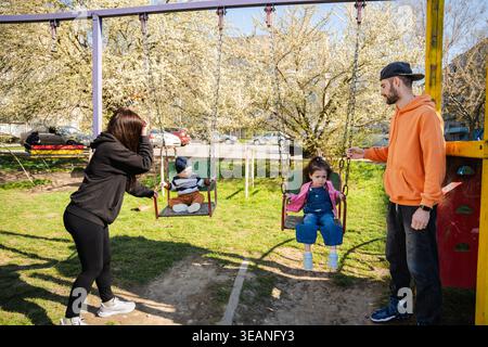Famiglia con due bambini seduti sulle altalene nel parco giochi con i genitori in piedi nelle vicinanze nel parco soleggiato. Concetto di tempo in famiglia e genitorialità. Foto Stock