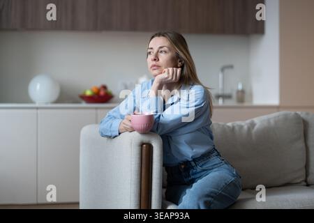 Una donna di mezza età leggermente triste seduta sul divano di casa con una tazza di caffè annoiata tenendo il mento con la mano Foto Stock