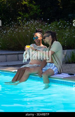 Coppia afroamericana che immerge i piedi nella piscina blu indossando una copertura all'uncinetto e tenendo un cocktail. Acqua, ponte, occhiali da sole, piastrelle, bevande, giardino, passante Foto Stock