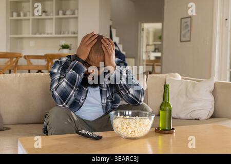Uomo adulto che indossa una camicia a quadri seduto nel soggiorno su un divano beige che regge la testa, ciotola di popcorn. Teso, ansioso, contemplativo, casa, interni, neutro, Foto Stock