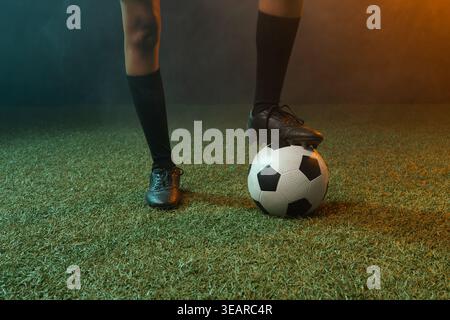 Donna afroamericana che bilancia il piede su una palla da calcio su erba in scarpe nere, calze al ginocchio. Atleta, equilibrio, allenamento, concentrazione, illuminazione, teal, amb Foto Stock