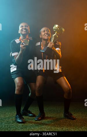 Donne afroamericane che festeggiano sul tappeto erboso, reggono il trofeo d'oro e la medaglia sotto le luci blu arancioni. Calcio, compagni di squadra, cameratismo, festeggiamenti, promo Foto Stock