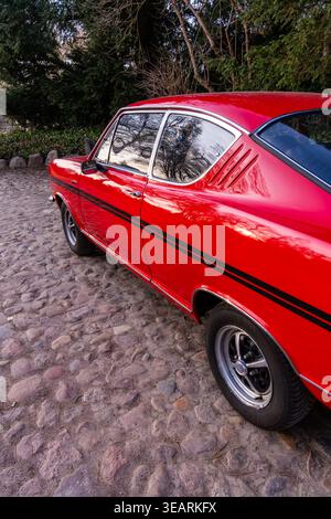 Una vivace coupé rossa d'epoca parcheggiata su una superficie rocciosa acciottolata, che mette in risalto il suo elegante profilo laterale e le finestre riflettenti. La striscia nera da corsa Foto Stock