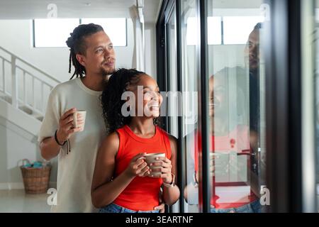 Coppia afroamericana in piedi davanti a porte di vetro che tengono le tazze donna che indossa la parte superiore rossa che guarda fuori. Intimo, sereno, accogliente, minimalista, moderno, giorno Foto Stock