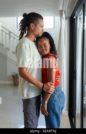 Coppia afroamericana in piedi che abbraccia all'ingresso da porte di vetro, tende a vista, cesto intrecciato. Romanticismo, coppia, abbraccio, casa, luce del giorno, finestra, scala Foto Stock
