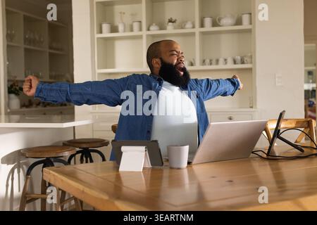 Uomo afro-americano che allarga le braccia e sbadiglia presso l'area di lavoro del tavolo da pranzo in legno con un computer portatile. Cucina, home Office, schermi, telecomando, scaffali, bar Foto Stock